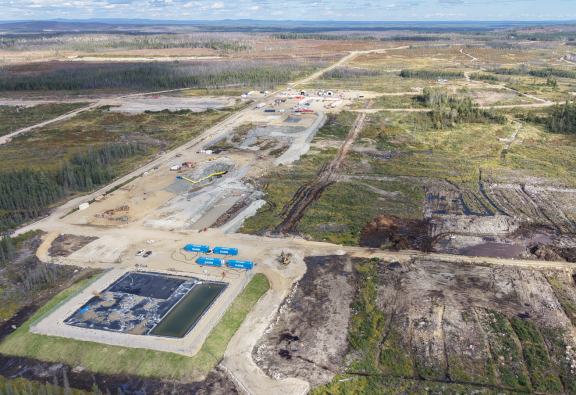 The image depicts a large industrial construction site with various machinery and equipment, surrounded by forested areas under a clear sky. Le contenu généré par l'IA peut être incorrect.