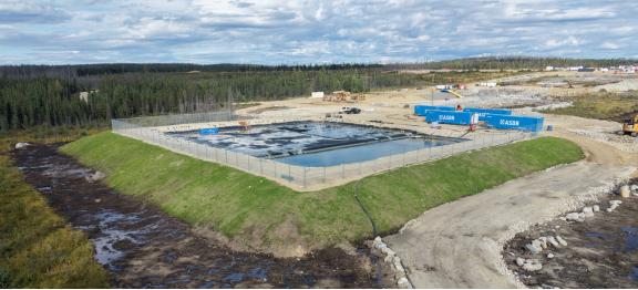 The image shows a construction site with a large, open area, surrounded by a chain-link fence, with machinery and equipment visible, under a clear blue sky with a few clouds. Le contenu généré par l'IA peut être incorrect.