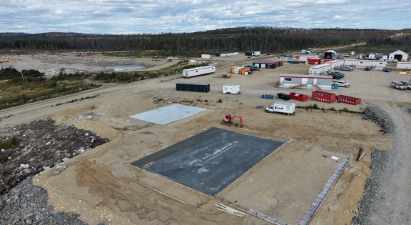 The image shows a construction site with several trailers, equipment, and a clear, unpaved area under a partly cloudy sky. Le contenu généré par l'IA peut être incorrect.