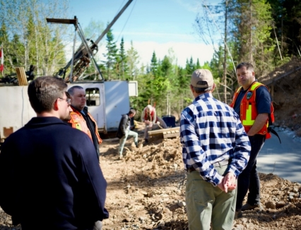 The image shows a construction site with workers, a crane, and a partially built structure surrounded by trees and a clear, cloudy sky.<br /> <br /> AI-generated content may be incorrect.