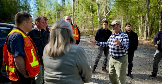 The image shows a group of people in safety vests and hats, gathered around a person holding a stick, in a wooded, dirt road environment.<br /> <br /> AI-generated content may be incorrect.