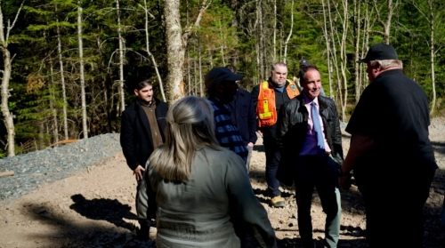 A group of people, some wearing winter clothing, stand in a forested area with a dirt path.<br /> <br /> AI-generated content may be incorrect.