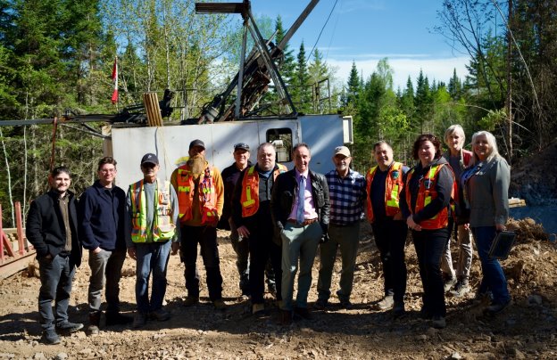 The image shows a group of construction workers, including a helmeted individual, gathered around a large, yellow crane, working in a dirt and rocky outdoor environment.<br /> <br /> AI-generated content may be incorrect.