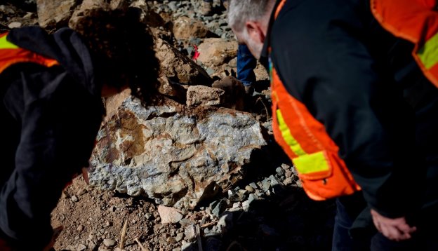 Two people are digging through a pile of rocky debris, wearing bright orange safety vests.<br /> <br /> AI-generated content may be incorrect.