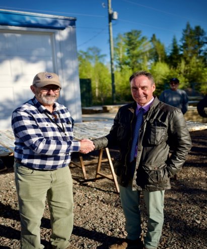 Two people shake hands in front of a blue building with a power pole in the background.<br /> <br /> AI-generated content may be incorrect.