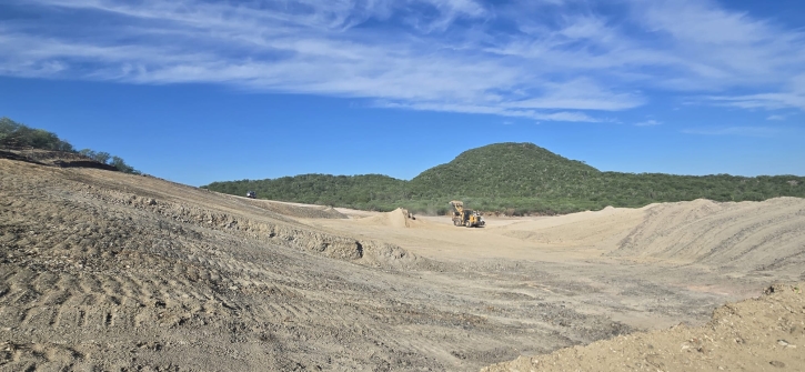 The image shows a large, barren, and dirt-filled excavation site with a mountainous background under a clear blue sky.<br /> <br /> AI-generated content may be incorrect.