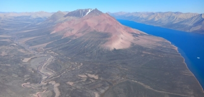 The image depicts a vast, barren, volcanic landscape with a prominent, red-colored mountain and a clear, expansive body of water in the background.<br /> <br /> AI-generated content may be incorrect.