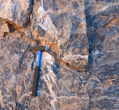 The image shows a black hammer leaning against a rough, textured rock surface.<br /> <br /> AI-generated content may be incorrect.