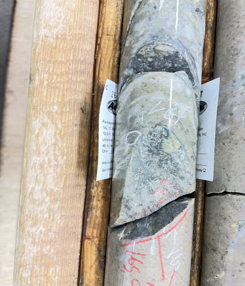 The image shows a wooden surface with a partially removed rock, displaying various labels and markings, including geochemical analysis and sample identification codes. AI-generated content may be incorrect.