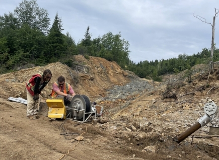 The image shows two people working on a construction site, using heavy machinery to excavate a dirt area under a cloudy sky, with trees in the background. AI-generated content may be incorrect.