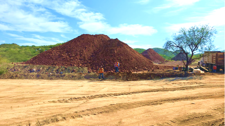 Workers are constructing a large pile of red dirt on an empty, grassy plot under a clear blue sky. AI-generated content may be incorrect.